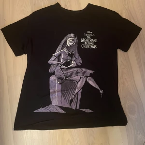 Svart Nightmare Before Christmas t-shirt - Svart t-shirt med tryck från Tim Burton's The Nightmare Before Christmas. Stor illustration av Sally som sitter med en svart katt, tryckt i lila och vitt. Rund halsringning och korta ärmar. Tröjan är köpt på Blue Fox i Stockholm, knappt använd. 