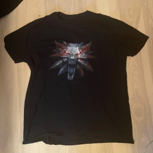 Svart Witcher t-shirt med tryck - Säljer en svart t-shirt med stort Witcher-tryck framtill. T-shirten har en klassisk passform och korta ärmar. Perfekt för dig som gillar gaming eller vill ha en cool statement-tee. Tröjan har tecken på användning på baksidan (stänkt från blekningsmedel) och trycket framtill är lite slitet. 