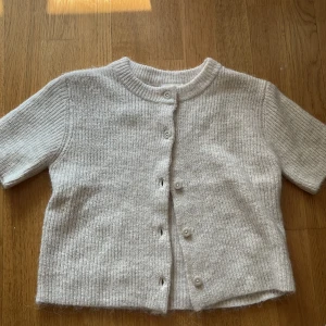 Beige stickad kofta från Gina Tricot - Säljer en beige stickad kofta från Gina Tricot med korta ärmar och rund hals. Koftan har ribbad struktur och knäpps med pärlemorknappar framtill. Perfekt att slänga över en topp för en chill look. Passar dig som gillar mjuka och neutrala färger.