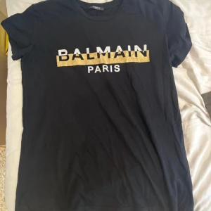 Svart Balmain Paris t-shirt i bomull - Svart t-shirt från Balmain Paris i mjuk bomull. Framsidan har en stor vit och guld Balmain Paris-logga med coolt tryck. Klassisk rund hals och korta ärmar, perfekt för dig som gillar streetstyle och designerplagg.