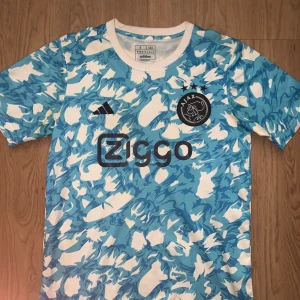 Ajax blå-vit kortärmad fotbollströja - Ajax Amsterdam fotbollströja från Adidas i slim fit. Tröjan har ett unikt blått och vitt mönster, korta ärmar och rund hals. Ajax-loggan med tre stjärnor på bröstet och Ziggo sponsortryck. Tillverkad i lätt, ventilerande polyester.