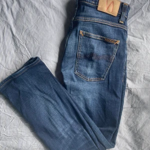 Blå jeans från Nudie Jeans Co - Snygga blå jeans från Nudie Jeans Co med klassisk femficksdesign och orangea kontrastsömmar. Jeansen har rak passform och är tillverkade i slitstarkt denim med coola detaljer som logotyp på bakfickan och läderpatch i midjan. Perfekta för dig som gillar stilrena och tidlösa jeans.