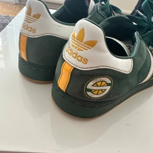 Adidas Superstar Seattle Sonics sneakers - Unika Adidas Superstar sneakers i Seattle Sonics NBA edition. Skorna har grön mocka, vita och gula detaljer, klassiska tre ränder på sidan och Sonics-logga på hälen och snörningen. Skalet framtill är beige och snörningen är grön. Perfekt för dig som gillar basket och retrostil ny pris 360dollar storlek 44