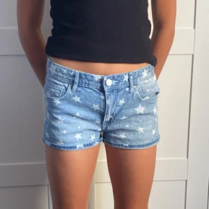 Blå jeansshorts med stjärnmönster - Snygga blå jeansshorts med vita stjärnor över hela tyget. Modellen är low waist/mid waist  och har klassiska fickor fram och bak samt knapp och dragkedja. Perfekta för sommarens alla häng, med en cool och ungdomlig vibe.