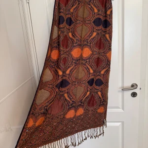 Mönstrad halsduk  - Snygg och stor halsduk med paisley-inspirerat mönster i brunt, orange och mörkblått. Tillverkad i 100% viskos, med glittriga detaljer och fransar längs kanterna. Perfekt att svepa runt halsen för en bohemisk vibe.