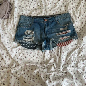 Blå jeansshorts med stjärnor och ränder - Snygga blå jeansshorts  med slitna detaljer, nitar vid fickorna och råa kanter. Insidan har coolt stjärnmönster och rödvita ränder som sticker ut under de trasiga partierna. Perfekt för dig som gillar statement-plagg och festivalvibe.