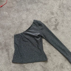 Glittrig oneshoulder topp Eight Paris - Säljer en svart glittrig topp från Eight Paris i storlek S. Modellen är oneshoulder med lång ärm på ena sidan och sitter tight mot kroppen. Materialet är mjukt och stretchigt, perfekt om du vill sticka ut med lite extra glitter. Supersnygg att matcha med jeans eller kjol.