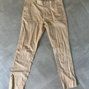 Beige chinos från COS, storlek 50 - Snygga beige chinos från COS i storlek 50. Byxorna har klassisk rak passform med bälteshällor och fickor både fram och bak. Tillverkade i 100% bomull för en skön och lätt känsla. Perfekta för en clean och stilren look. 1A5