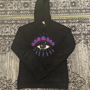 Kenzo Eye hoodie - Svart hoodie från Kenzo i storlek XS med ett stort broderat ögonmotiv i blått, lila och rosa på bröstet. Tröjan har huva med snörning och långa ärmar. Perfekt för dig som gillar statement-plagg och coola designer.