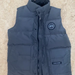 Mörkgrå dunväst från Canada Goose - Snygg mörkgrå dunväst från Canada Goose med hög krage, två stora fickor framtill och diskret logga på bröstet. Västen har dragkedja och tryckknappar samt klassisk quiltad design. Perfekt för lager-på-lager under kyliga dagar.