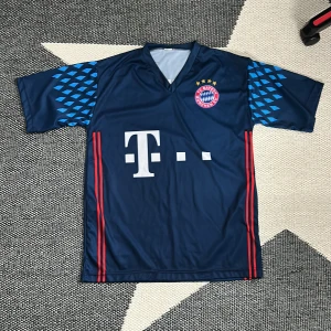 Bayern München Neuer fotbollströja - Snygg marinblå Bayern München fotbollströja med Neuer och nummer 1 på ryggen. Tröjan har korta ärmar med blå geometriskt mönster, röda ränder längs sidorna och klubbmärke med fyra stjärnor på bröstet. Tillverkad i lätt polyester, perfekt för match eller träning.