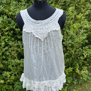 Vit topp med spets och volang - Supersöt vit topp  med bred spetsdetalj framtill och dekorativ kant vid halsen. Toppen är ärmlös och har knappar i ryggen samt volang längst ner. Perfekt för dig som gillar romantisk och boho-inspirerad stil. Trf Zara.