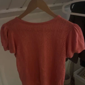 Korallrosa blus från JDY med puffärm - Superfin korallrosa blus från JDY i storlek M. Blusen har korta puffärmar och ett snyggt stickat hålmönster över hela plagget. Rund halsringning och mjukt material som känns skönt mot huden. Perfekt för dig som gillar romantiska detaljer och färg!
