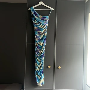 Blommig långklänning med oneshoulder - Säljer en superfin långklänning med blommigt mönster i blått, lila och beige. Klänningen har oneshoulder-design med en axelrem och draperade detaljer längs hela klänningen. Sitter tight och har en snygg passform som framhäver figuren. Perfekt för dig som vill sticka ut!