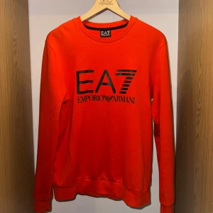 Orange EA7 Emporio Armani tröja - Snygg orange tröja från EA7 Emporio Armani med svart logotyptryck på bröstet. Rund halsringning och långa ärmar. Mjuk och skön bomullskvalitet som passar perfekt till streetwear. Enkel design med ribbade muddar och klassisk passform.