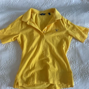 Gul piké t-shirt från GANT, S - Fräsch gul piké t-shirt från GANT i storlek S. Klassisk krage, korta ärmar och broderad logga på. 4 knappar vid halsen. Perfekt för dig som gillar sportig och clean stil.