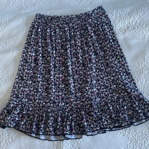 Blommig kjol från Ü - Supersöt kjol från Ü med svart botten och blommigt mönster i vitt och rosa. Klänningen har volangkant nedtill och ett elastiskt midjeband för skön passform. 