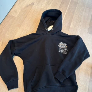 Svart Stüssy hoodie med tryck - Svart hoodie från Stüssy med stort grafiskt tryck och text på ryggen samt mindre tryck på bröstet. Klassisk huva med metallöljetter och känguruficka framtill. Mjuk bomullskvalitet och ribbade muddar vid ärmar och nederkant. Perfekt för streetwear-stilen.
