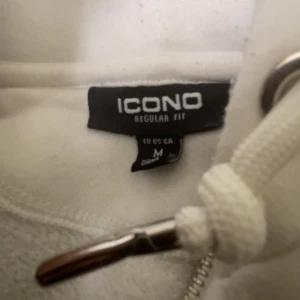 Vit hoodie från ICONO med dragkedja - Säljer en fräsch vit hoodie från ICONO i storlek M. Tröjan har huva med snörning, dragkedja framtill och coolt märke på bröstet. Materialet är mjukt och skönt, med paneler i ljusbeige och snygga detaljer längs dragkedjan. Perfekt för chill eller streetwear.