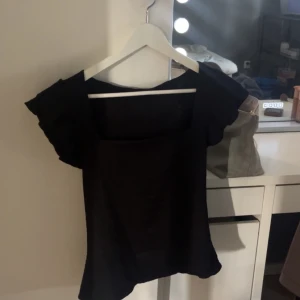 Svart blus med volangärmar - Snygg svart blus med fyrkantig urringning och korta volangärmar. Perfekt för dig som gillar en enkel men trendig look. Materialet känns lätt och luftigt, och toppen har en loose passform som ger en chill vibe.