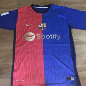 FC Barcelona Lamine Yamal tröja Nike - Säljer en FC Barcelona matchtröja med Lamine Yamal och nummer 19 på ryggen. Tröjan är kortärmad och har klassisk halva-röd halva-blå design med guldiga detaljer och Spotify-logga på bröstet. Materialet är lätt och andas, perfekt för fotboll eller häng. Officiell Nike Dri-FIT ADV.