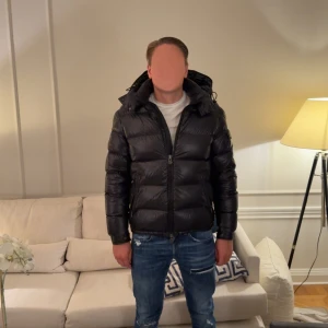 Moncler Maya giboutto - Svart pufferjacka från Moncler med glansig finish, stor huva och Moncler-logga på ärmen. Jackan har dragkedja framtill, fickor med dragkedja och en extra ficka med knapp på ärmen. Passformen är normal och jackan är perfekt för kalla dagar. Kvitto finns, köpt den personligen i butik här i Stockholm för 2 år sedan. Storlek 3/L