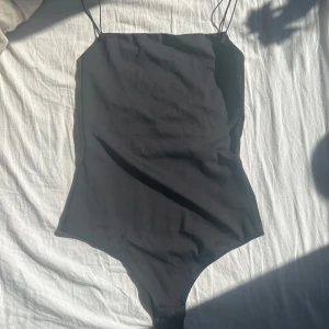 Svart ärmlös bodysuit med smala band - Svart bodysuit med smala axelband och rak halsringning. Sitter tight och har en clean, minimalistisk look. Perfekt att styla med jeans eller kjol för en cool vibe. Materialet känns mjukt och stretchigt, vilket gör den bekväm att bära hela dagen.