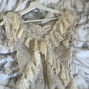 Beige spetsblus från Zara - Supersöt beige blus från Zara med breda volanger i spets och stickad struktur. Ärmlös modell med v-ringad hals och detaljerad broderad spets framtill och runt axlarna. Perfekt för dig som gillar romantisk och boho-inspirerad stil.de ca långärmad fört men har klippt bort dom men fortfarande lika fin