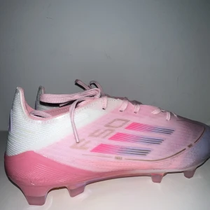 Adidas rosa fotbollsskor FSO - Säljer ett par Adidas FSO fotbollsskor i en snygg rosa och vit färg med lila detaljer. Skorna har snörning och en platt sula med dobbar i iriserande finish. Ovandelen är tillverkad i syntetmaterial och har de klassiska tre ränderna i rosa och blått på sidan. Perfekt för dig som vill sticka ut på planen.