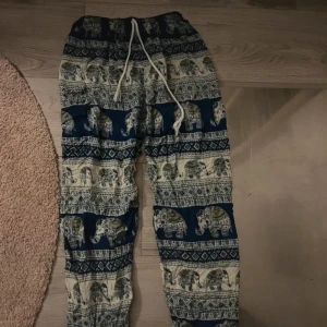 Blå linnebyxor med elefantmönster - Säljer ett par chill blå linnebyxor med unikt elefantmönster och vita snören i midjan. Byxorna har en loose passform och är perfekta för dig som gillar boho-vibe och sköna material. Mönstret är detaljerat med inslag av grönt och gult på elefanterna.