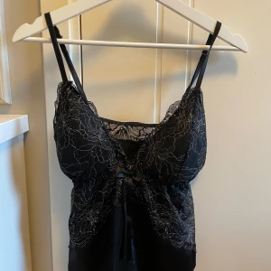 Svart spetstopp med rosett - Säljer en svart topp med tunna axelband och v-ringning. Toppen har ett blommigt spetsmönster och en liten rosett framtill. Materialet är mjukt och glansigt, troligen satin eller polyester, vilket ger en lyxig känsla. Perfekt för dig som gillar detaljer och en feminin look. Storlek L men sitter som XS-M.