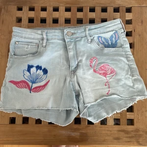 Broderade ljusblå jeansshorts H&M - Unika ljusblå jeansshorts från H&M x Michelle Morin med broderade detaljer som flamingo, fjäril och blomma i rosa, blått och vitt. Shorts med rå kant, fem fickor och dragkedja. Perfekt för dig som vill sticka ut med coola broderier.