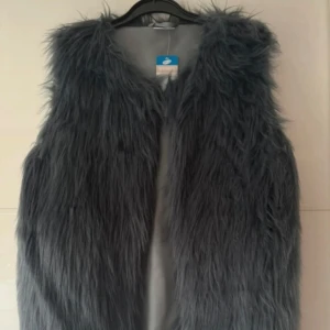 Ny, grå långhårig pälsväst från Below - Säljer en snygg grå pälsväst från Below i storlek M. Västen har lång, fluffig fuskpäls och är helfodrad med slät grå insida. Stängs med diskret hyska framtill. Perfekt för lager-på-lager och ger en cool vibe till din outfit.