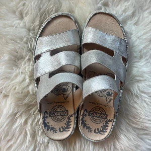 Silvriga sandaler från Brako, strl 37 - Säljer ett par sandaler från Brako i storlek 37. De har tre breda remmar i skimrande silverfärgat skinn och en platt, vit sula med mönstrad kant. Innersulan har ett blommigt tryck och är mjuk för extra komfort. Perfekta för dig som gillar glittriga detaljer och snygg design. Helt nya💓💓