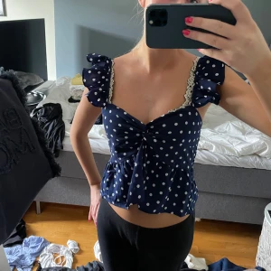 Mörkblå prickig blus med volang - Supersöt mörkblå blus med vita prickar och volangdetaljer på axelbanden. Blusen har en twistad front, spetskant vid axelbanden och är croppad med en luftig passform. Perfekt för dig som gillar romantisk och lekfull stil.