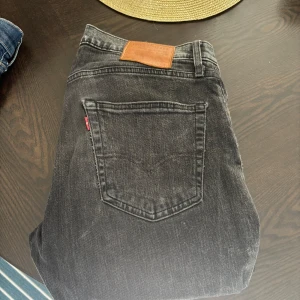 Levi's 502 svarta jeans W31 L32 - Svarta Levi's 502 jeans med klassisk femficksdesign och brunt läderpatch bak i midjan. Jeansen har en rak passform och är tillverkade i slitstarkt denim med en cool, något tvättad look. Perfekta för dig som gillar tidlös streetstil och vill ha ett par jeans som funkar till allt.