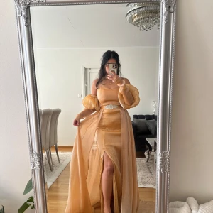 Champagnefärgad långklänning med puffärmar - Säljer en elegant champagnefärgad långklänning med transparenta puffärmar och slits framtill. Klänningen har omlott-detalj i kjolen och ett dekorativt bälte med pärlor i midjan. Materialet är lätt och skimrande, perfekt för bal eller fest. Passformen är normal och ärmarna är långärmade. Har köpte och ej använd den klänningen finns kvar kvitto 