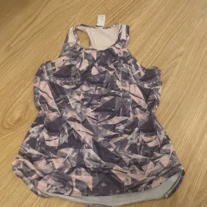 Mönstrad träningstopp i lila och rosa - Säljer en sportig linnetopp med coolt grafiskt mönster i lila och ljusrosa toner. Toppen har brottarrygg och ärmlös design, perfekt för gymmet eller löprundan. Materialet är lätt och andas, vilket gör den skön att bära under träning.