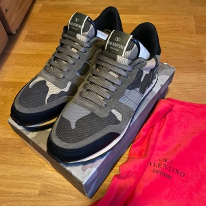 Valentino Garavani camo sneakers strl 42 - Snygga Valentino Garavani Rockrunner sneakers i grått och svart med camouflage-mönster. Skorna har mesh och mocka-paneler, snörning och en cool gummisula med pyramidmönster. Perfekt för dig som vill sticka ut med exklusiva och trendiga sneakers.