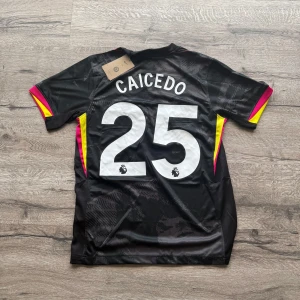 Chelsea Caicedo #25 svart matchtröja - Chelsea FC Caicedo #25 svart fotbollströja från Nike med korta ärmar. Tröjan har rosa och gula detaljer på ärmarna, ett subtilt mönster över hela tyget och klubbens lejonlogga på bröstet. Materialet är lätt och andas, perfekt för träning eller match. Storlek: S helt ny med prislapp. Priset är inte hugget i sten så skicka pm vid funderingar.