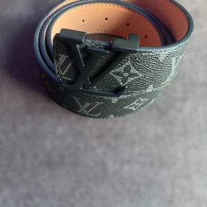Louis Vuitton svart skinnbälte - Snyggt svart bälte från Louis Vuitton med klassiskt monogrammönster och stor LV-logga i mattsvart metall. Bältet är gjort i präglat skinn med beige insida och har en robust spänne med Louis Vuitton-gravyr. Perfekt för dig som vill ha en lyxig och trendig accessoar.