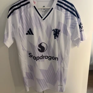 Manchester United Cunha 10 Adidas tröja S - Säljer en Manchester United fotbollströja med Cunha 10 på ryggen. Adidas slim fit med V-ringning, korta ärmar och lila mönster på vit bakgrund. Tre svarta ränder på axlarna, klubbmärke och Snapdragon-logga framtill. Tillverkad i lätt polyester för maximal komfort. Byte funkar också mot andra nya tröjor! 