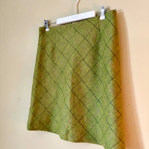 Grön rutig ullkjol från Newhouse - Supersnygg grön kortkjol från Newhouse i 100% ull med rutigt mönster i mörkgrönt och rosa. Kjolen har dragkedja bak och är helfodrad. Perfekt för dig som gillar klassisk stil med en twist!