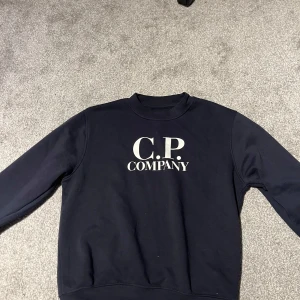Mörkblå sweatshirt från C.P. Company - Snygg mörkblå sweatshirt från C.P. Company i storlek S. Tröjan har ett klassiskt crewneck-snitt och stor vit logga på bröstet. Tillverkad i mjuk bomull med långa ärmar och ribbade muddar vid ärmslut och nederkant. Perfekt för en chill och stilren look.