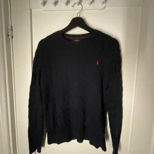 Mörkblå stickad tröja Polo Ralph Lauren - Snygg mörkblå stickad tröja från Polo Ralph Lauren i storlek 18-20 som motsvarar ungefär storlek S. Tröjan har ett klassiskt kabelstickat mönster och en liten röd broderad logga på bröstet. Rund hals och långa ärmar, perfekt för dig som gillar stilren och tidlös design.