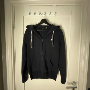 Mörkblå hoodie från Polo Ralph Lauren - Snygg mörkblå hoodie från Polo Ralph Lauren med vit snörning i huvan och dragkedja framtill. Klassisk logga broderad på bröstet och två fickor. Perfekt för chill dagar och enkel att matcha med jeans eller joggers.