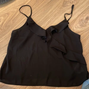Svart linnetopp med volang från Gina Tricot - Svart linnetopp från Gina Tricot i storlek 36. Tunn och flowy polyester med justerbara smala axelband och snygg volangdetalj framtill. Perfekt att styla med jeans eller kjol för en chill och trendig look.