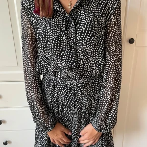 Svartvit prickig kortklänning från Zara - Säljer en svartvit kortklänning från Zara med prickigt mönster. Klänningen har långärmade transparenta ärmar, knappar framtill och ett knytband i midjan för snygg passform. Materialet är lätt och luftigt, perfekt för att sticka ut med coola detaljer.