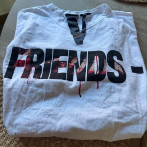 Vit VLONE Friends t-shirt XL - Säljer en vit t-shirt från VLONE i storlek XL med stor svart och röd 'FRIENDS-' print på framsidan och ett stort V med blodstänk på ryggen. T-shirten har rund hals och är kortärmad, tillverkad i mjuk bomull. Perfekt för dig som gillar streetwear med edge. Aldrig använd pga för liten storlek, nypris 3900:- säljes för 2000:-