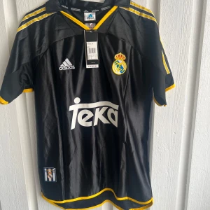 Real Madrid svart Raul 7 Adidas tröja - Snygg svart Real Madrid fotbollströja från Adidas med gula detaljer och klassiska ränder på axlarna. Tryck med Teka på bröstet och Raul 7 på ryggen. Broderat klubbmärke på framsidan. Tillverkad i lätt polyester som andas.